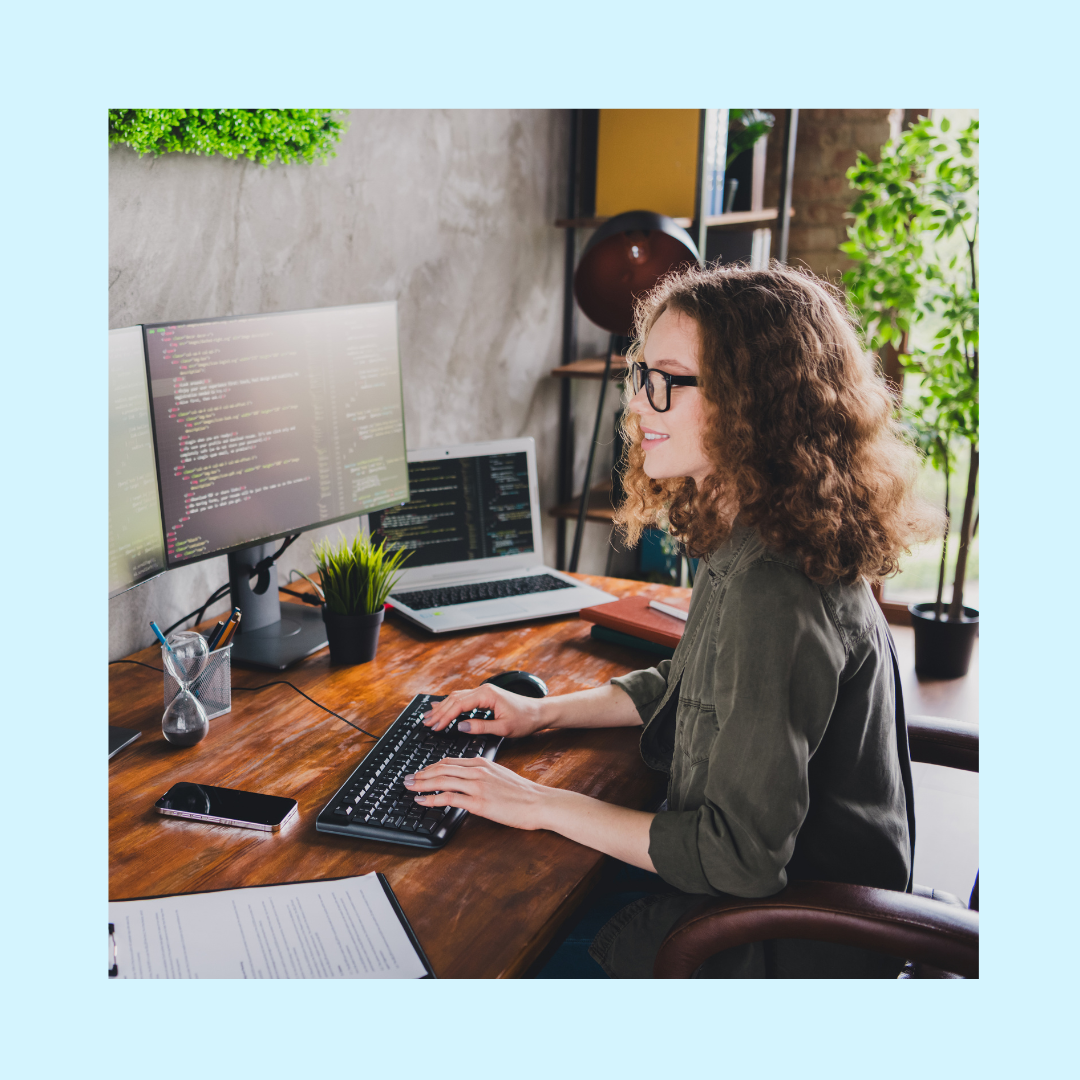 Woman with curly hair and glasses working at a wooden desk, typing on a keyboard while looking at dual monitors displaying lines of code, with a laptop, smartphone, notebook, and small plants arranged in a modern, well-lit workspace.