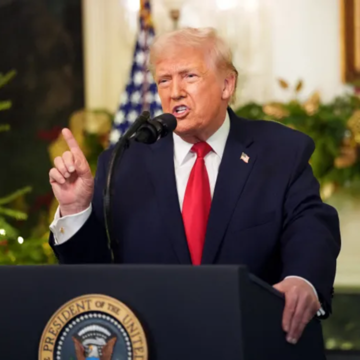 President Trump speaking at a podium in the White House, wearing a navy suit and red tie