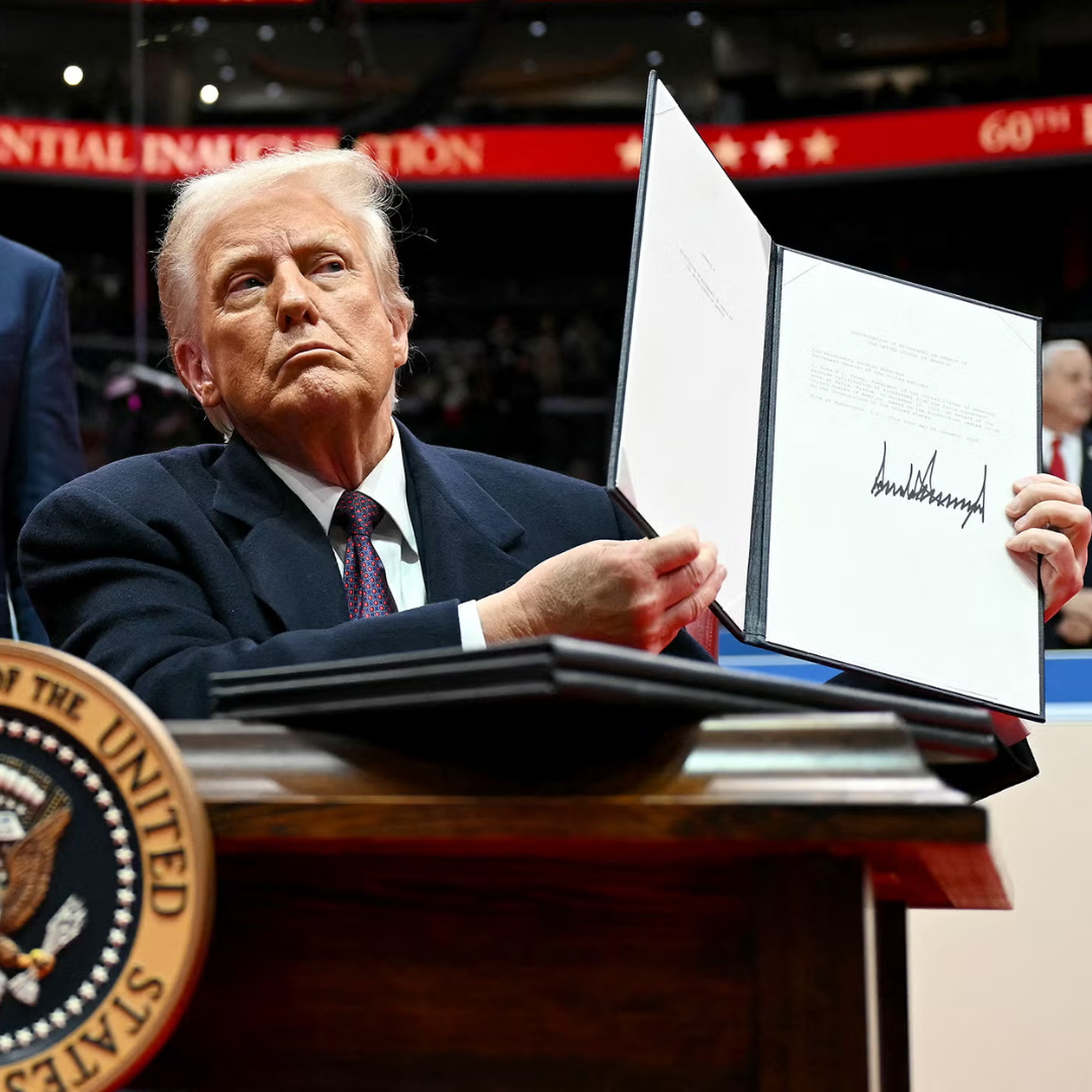 President Donald Trump holding up a signed document while seated at a desk with the U.S. presidential seal visible