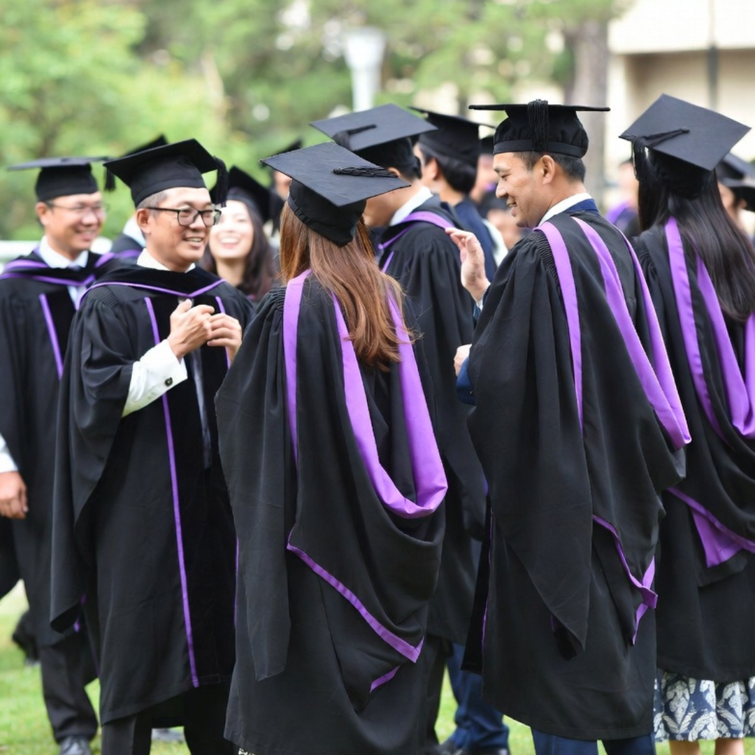 A group of graduates wearing black caps and gowns with purple accents stand outdoors at a graduation ceremony