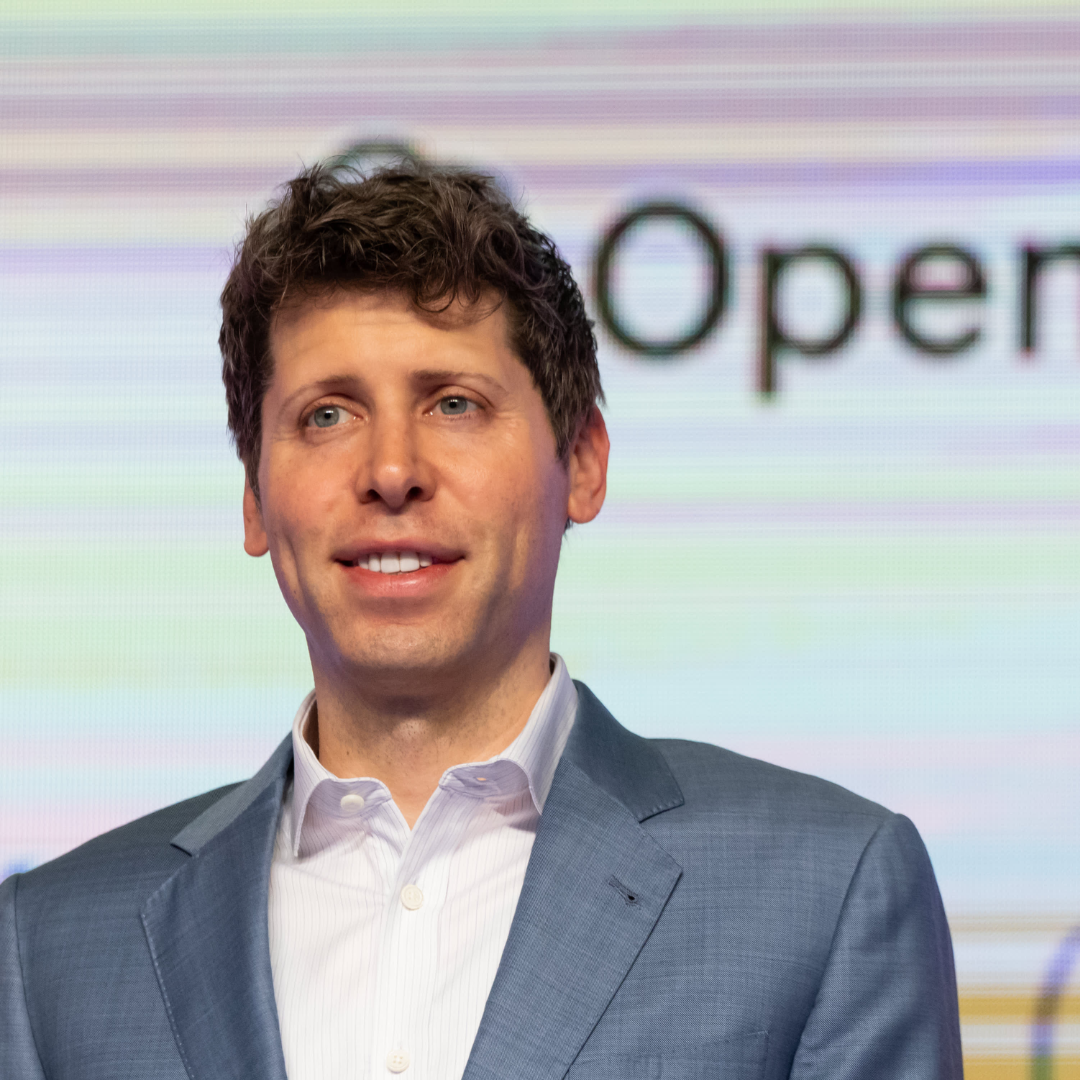 OpenAI CEO Sam Altman speaking at an event, with the OpenAI logo blurred in the background.