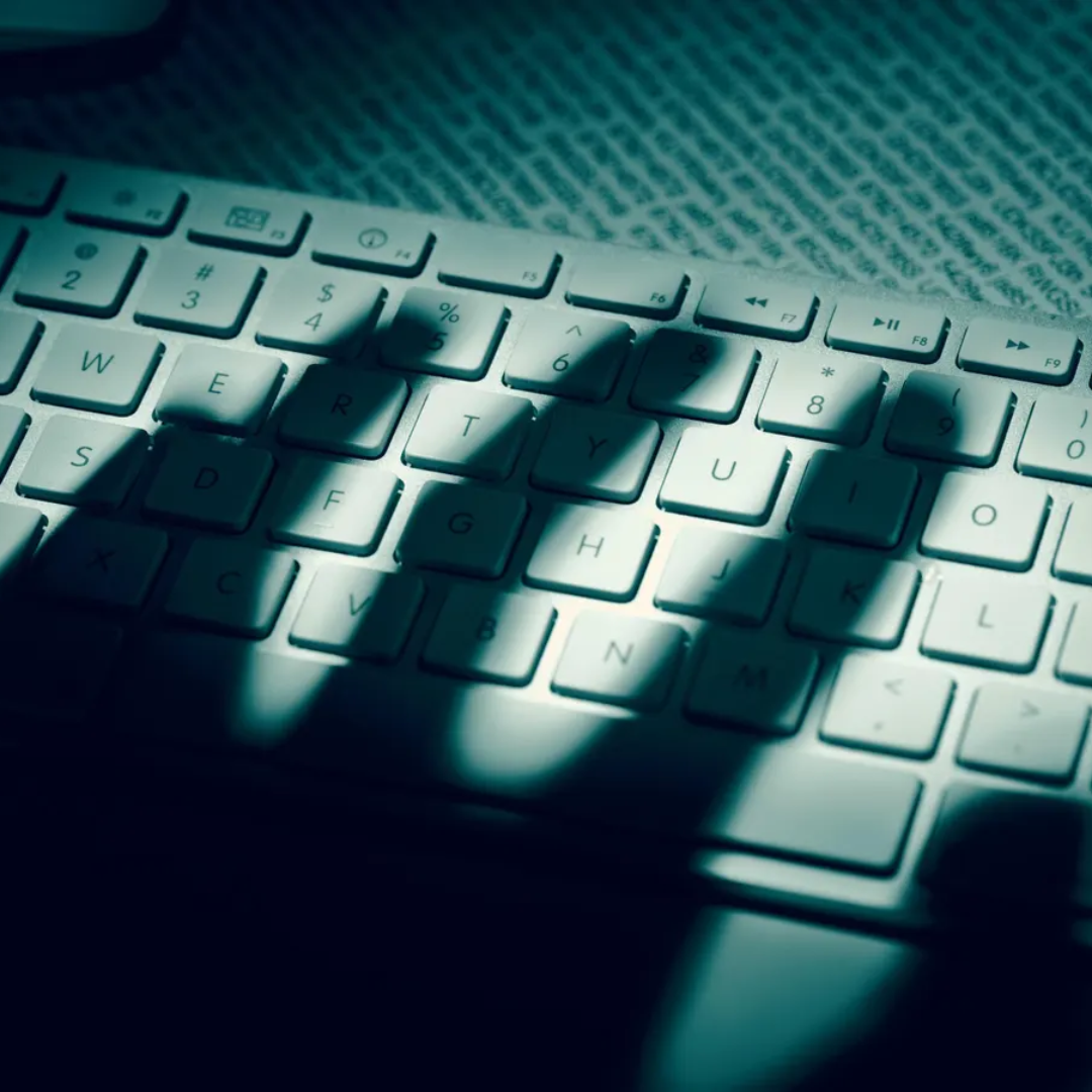 Close-up of a computer keyboard in dim, shadowy lighting with a teal tint and a hand's shadow cast across the keys