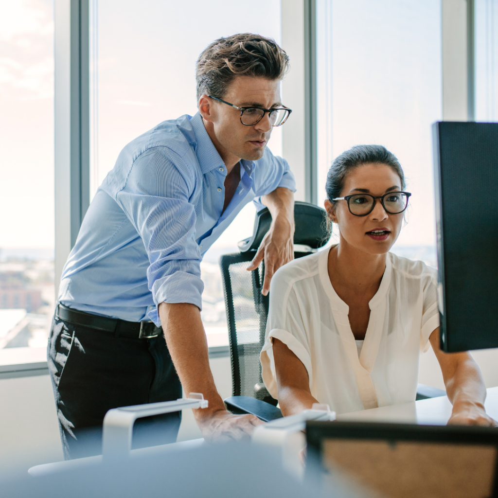 Two administrative colleagues wearing glasses collaborating at a computer in a bright office with large windows