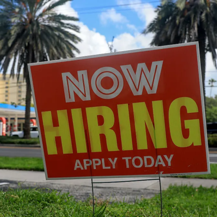 Red "Now Hiring Apply Today" yard sign in grass with palm trees in the background