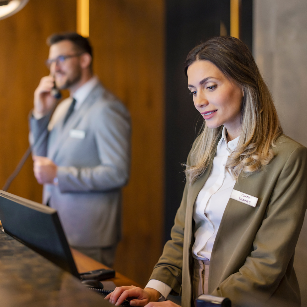 Two hotel front desk staff members in uniform working at a reception counter, one on the phone and one at a computer