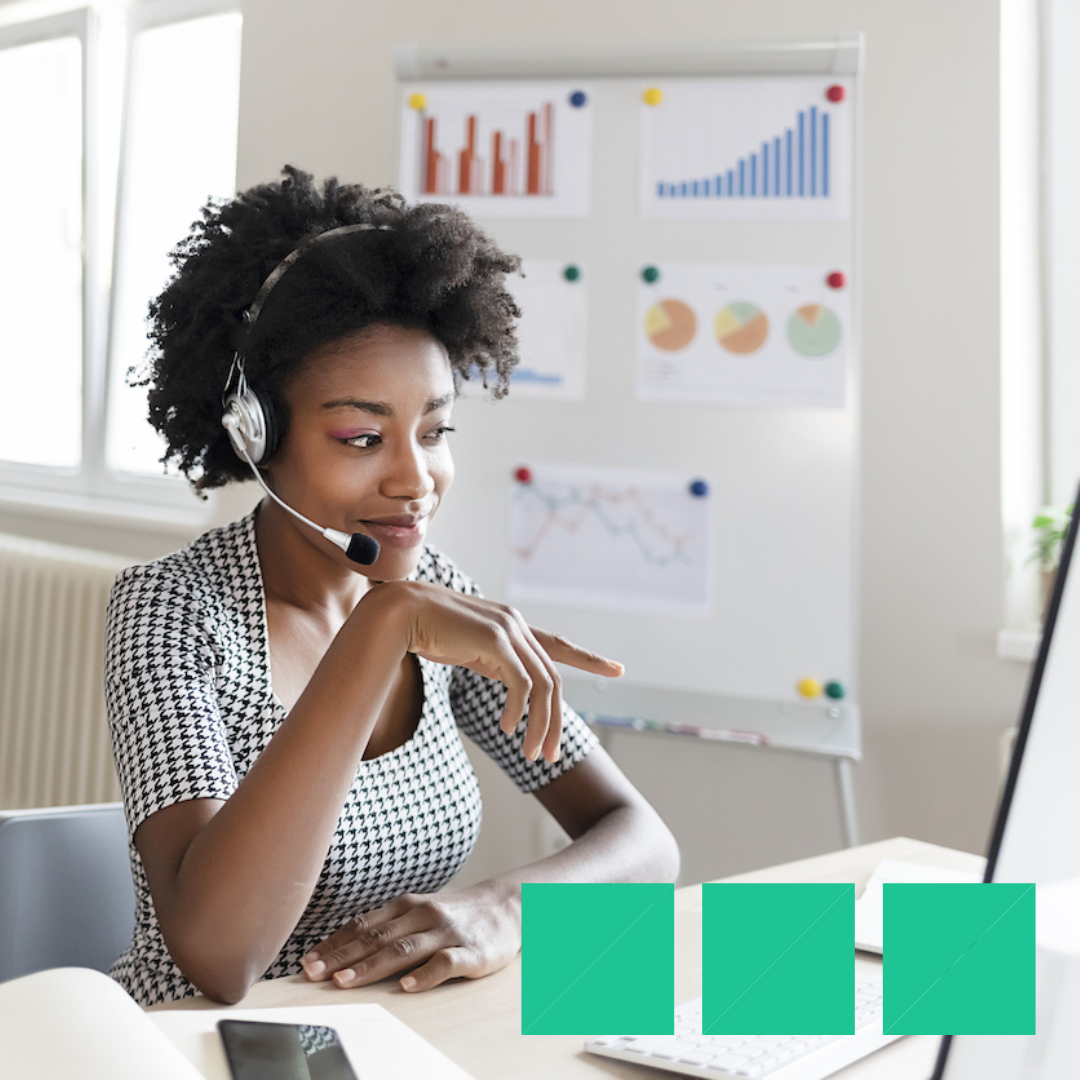 Woman wearing a headset at a desk looking at a computer monitor with business charts on a whiteboard in the background