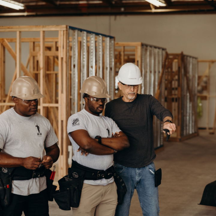 Construction supervisor in a white hard hat directing two workers wearing tan hard hats inside a building under construction
