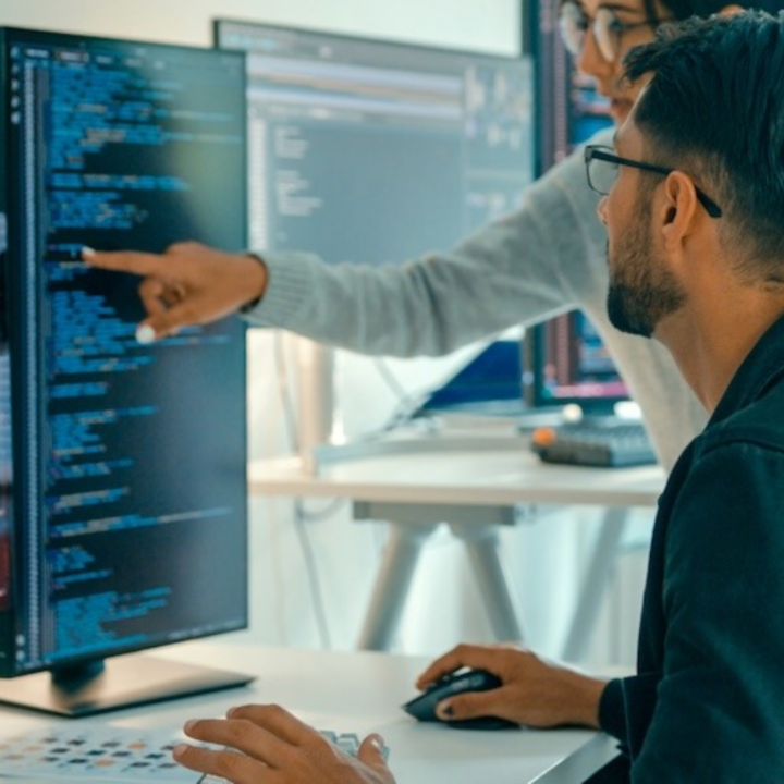 Two employees collaborating at a workstation with multiple monitors displaying code