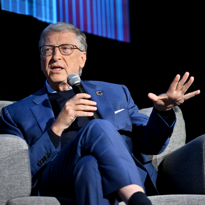 Bill Gates speaking on stage with a microphone while seated, wearing a blue suit