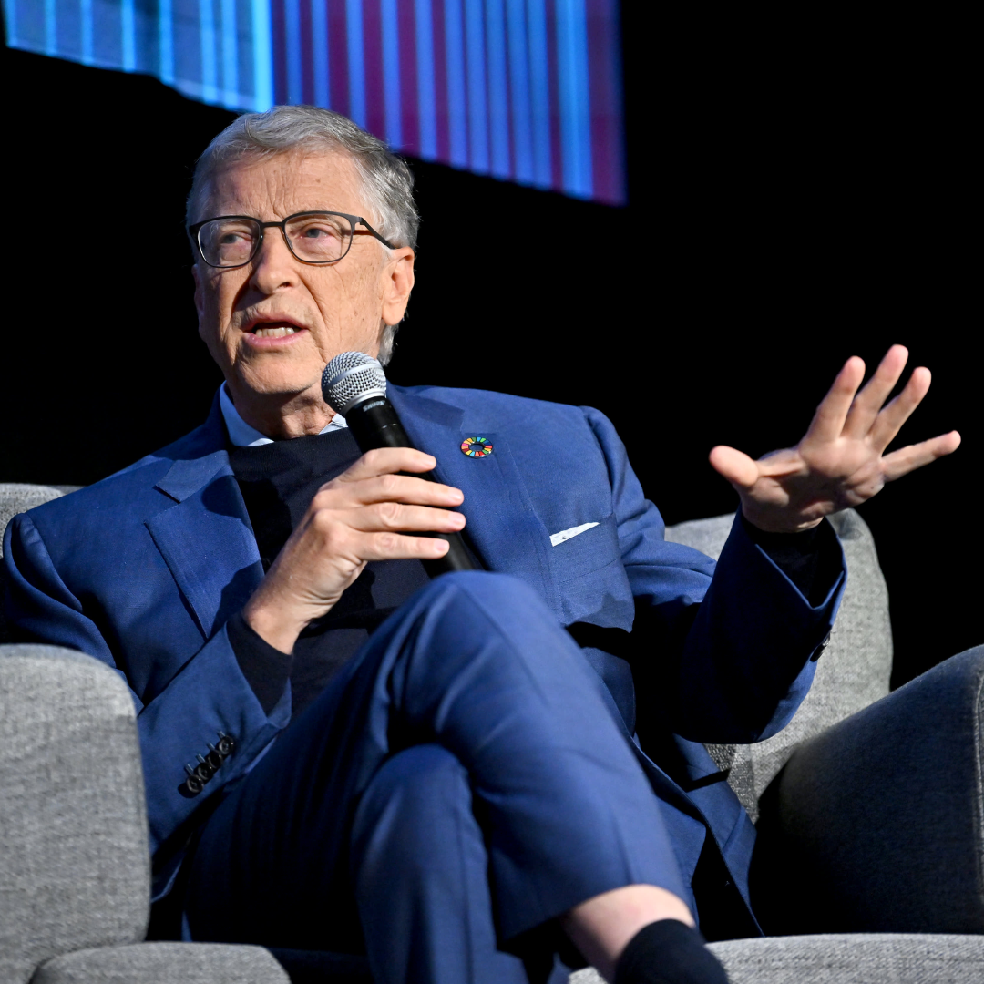 Bill Gates speaking on stage with a microphone while seated, wearing a blue suit