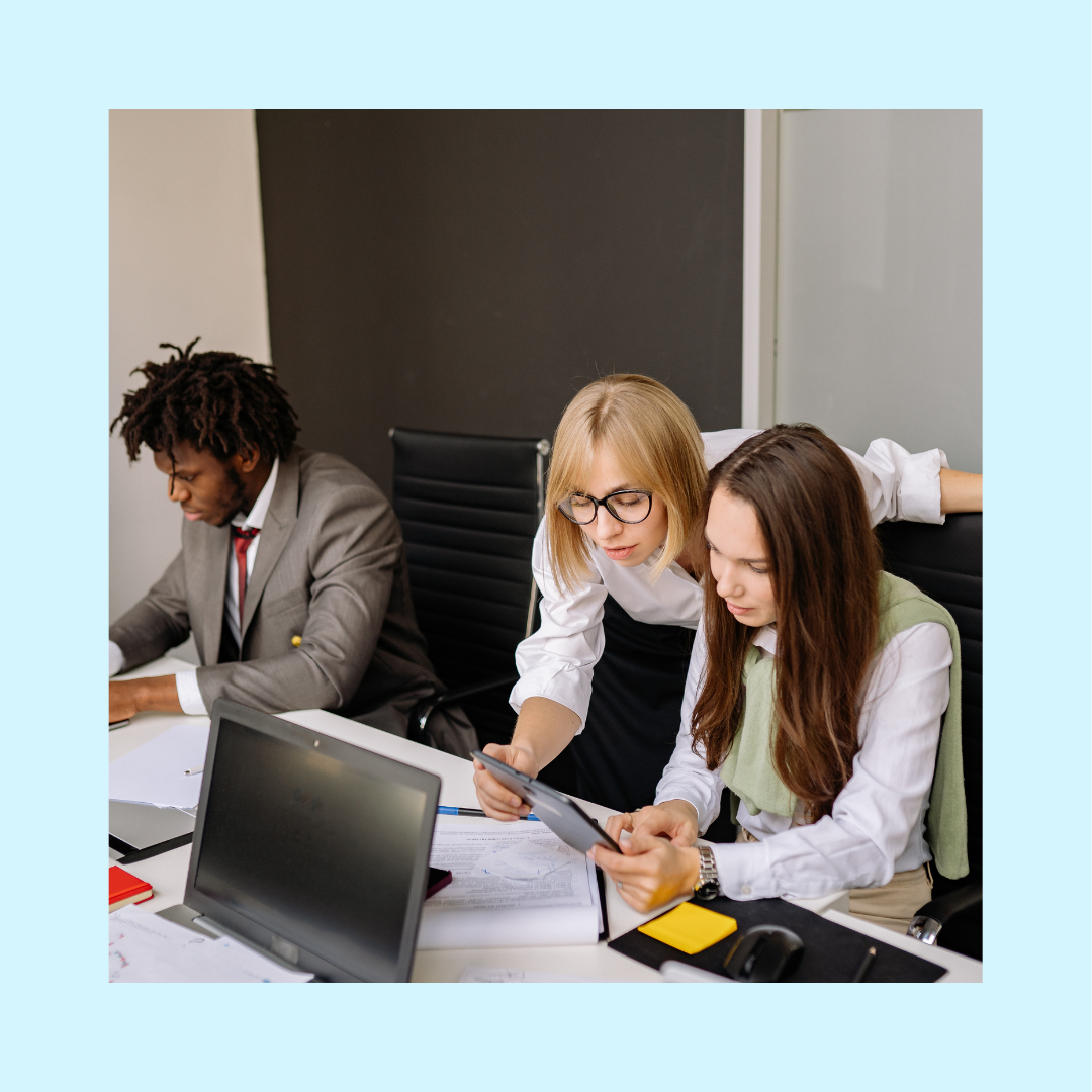 Three office professionals working at a conference table, with two women reviewing documents together and a man working independently nearby