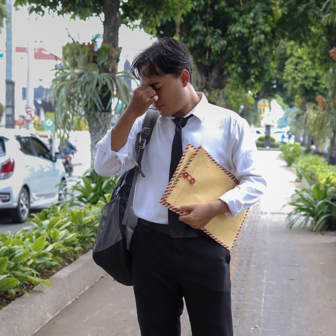 Stressed young man in a dress shirt and tie holding a manila envelope and backpack on a city sidewalk