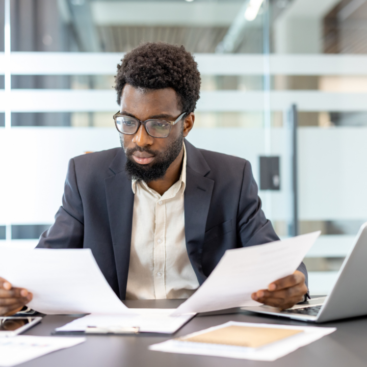 Businessman wearing glasses reviewing documents at a desk with a laptop in a modern glass-walled office