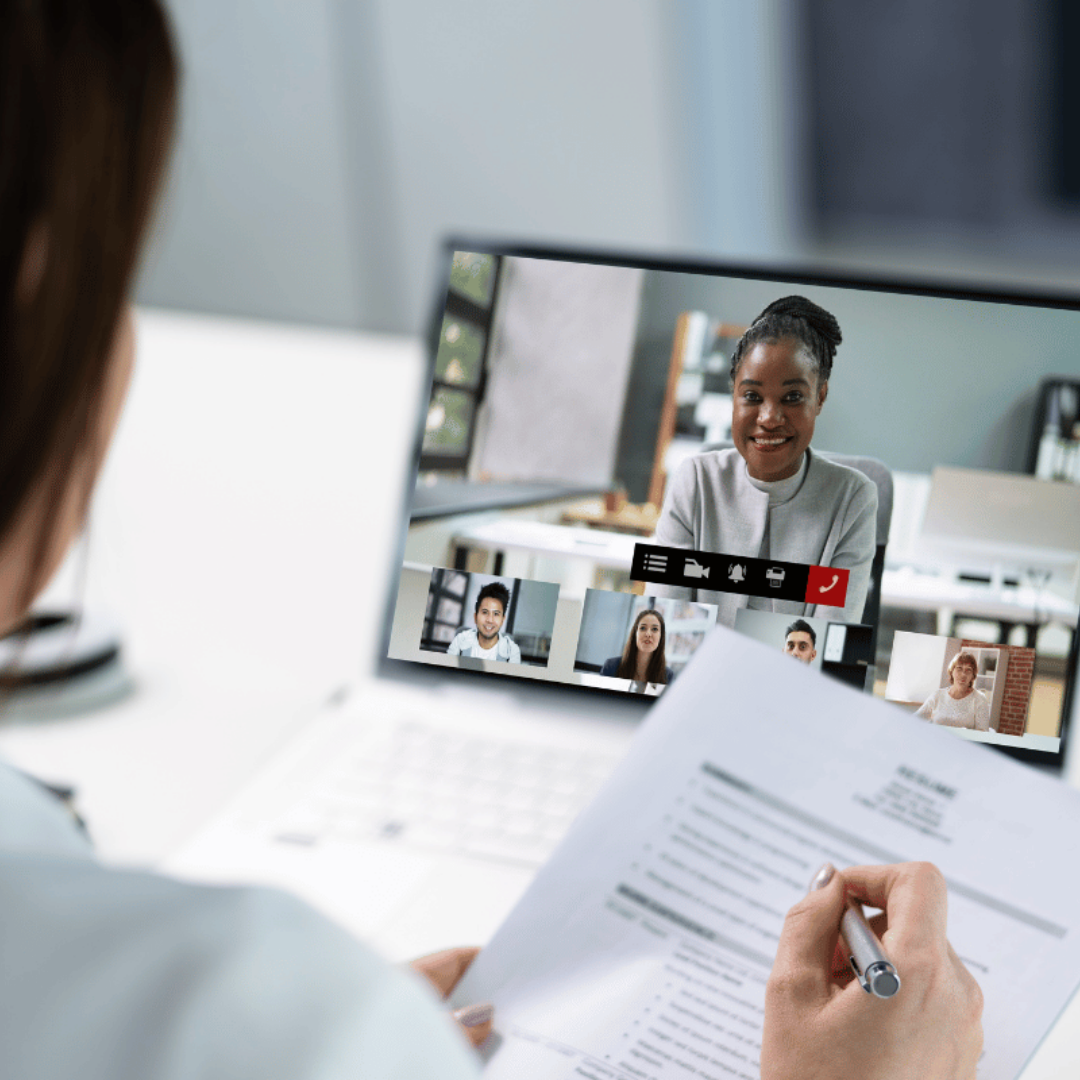 Woman holding a resume during a video job interview with multiple participants visible on a laptop screen