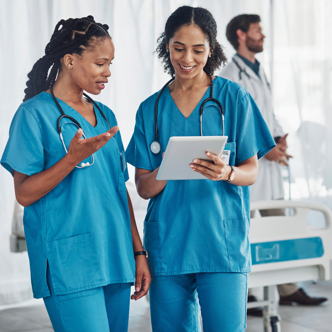 Two nurses in blue scrubs reviewing information on a tablet in a hospital room with a physician visible in the background