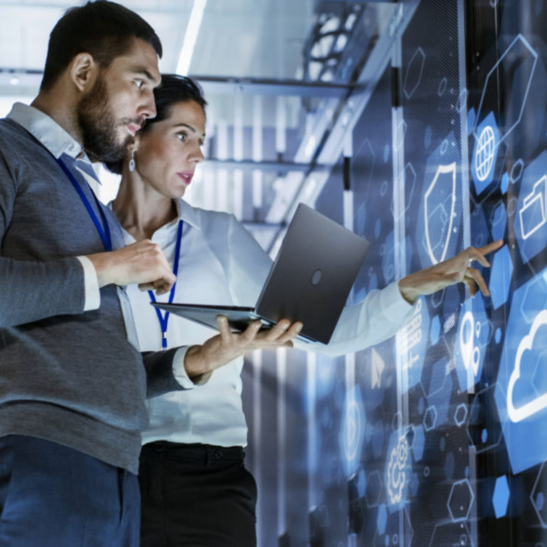 Two IT professionals with a laptop examining a digital display showing cybersecurity and cloud computing icons in a server room
