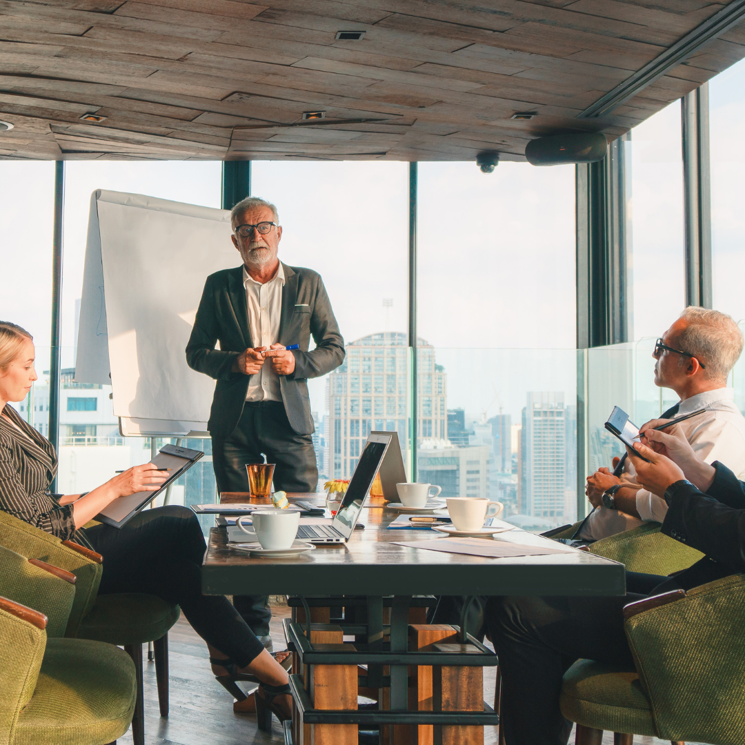 CEO presenting to colleagues in a high-rise conference room with a city skyline view and whiteboard behind him