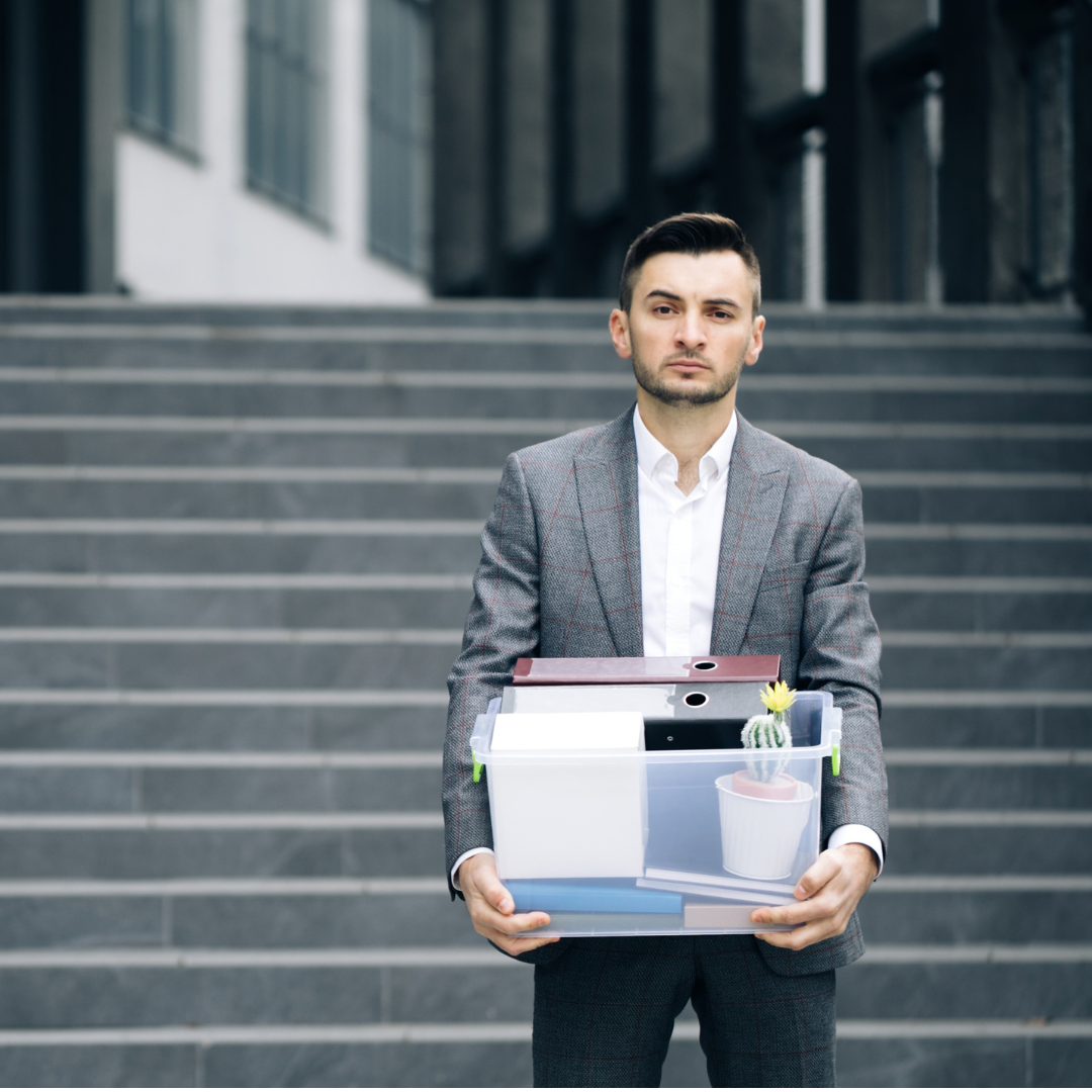 Man in a suit standing on office building steps holding a clear plastic bin with personal belongings including a small cactus plant