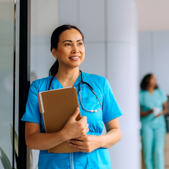 Smiling nurse in blue scrubs holding a folder with a stethoscope around her neck in a hospital hallway