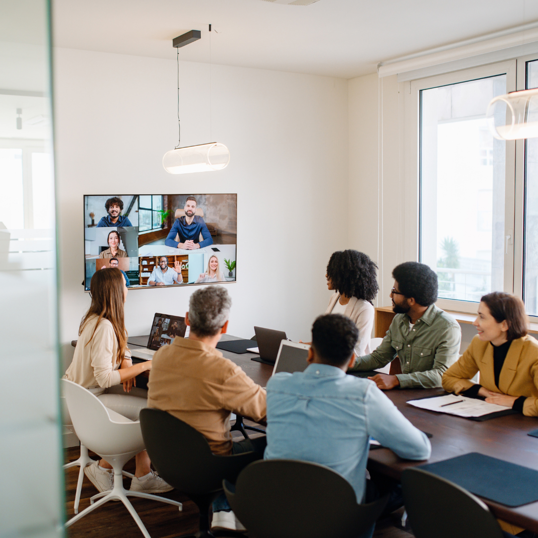 Diverse team of employees in a conference room participating in a hybrid meeting with remote colleagues visible on a wall-mounted screen