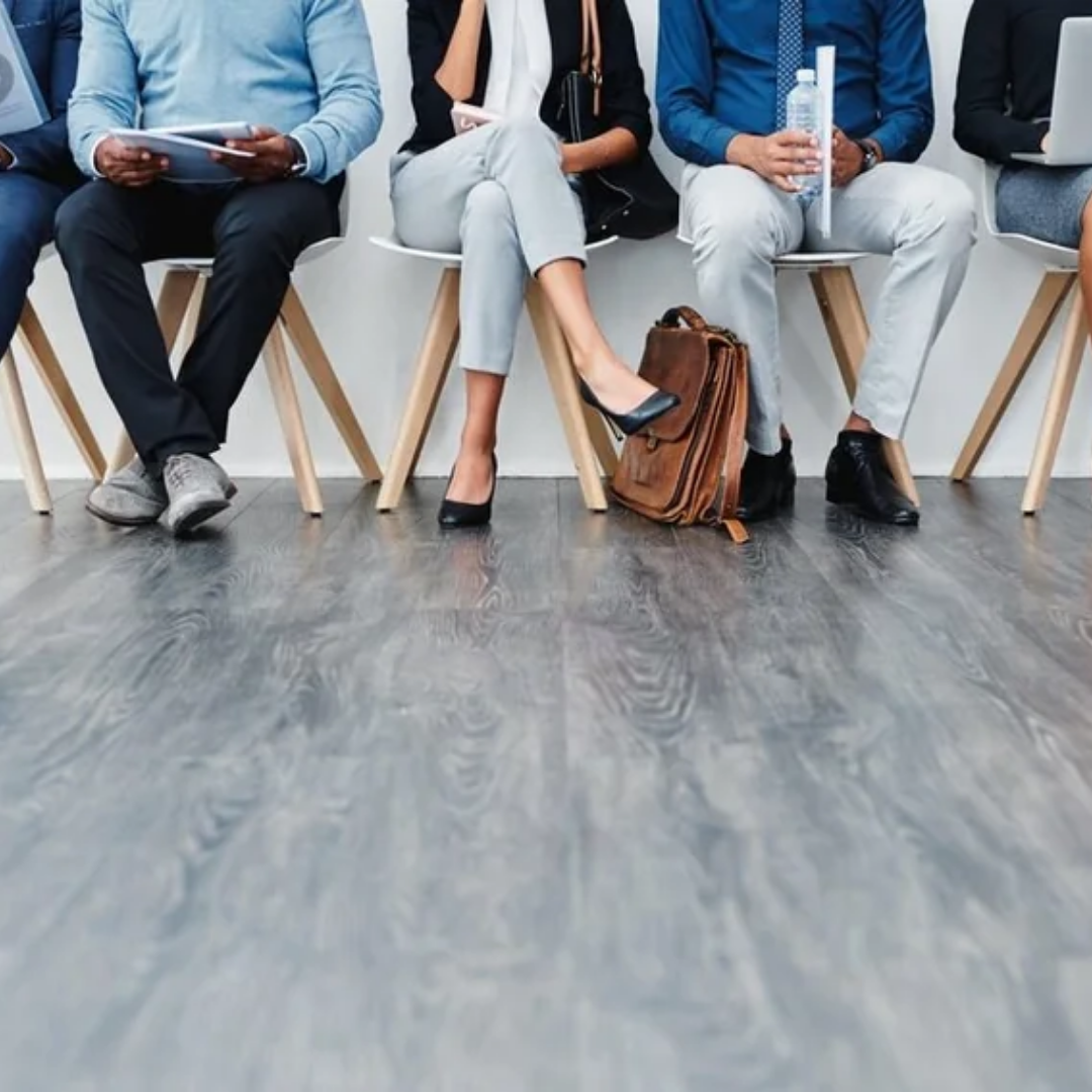 Row of job candidates seated in a waiting area holding documents and devices, shown from the waist down