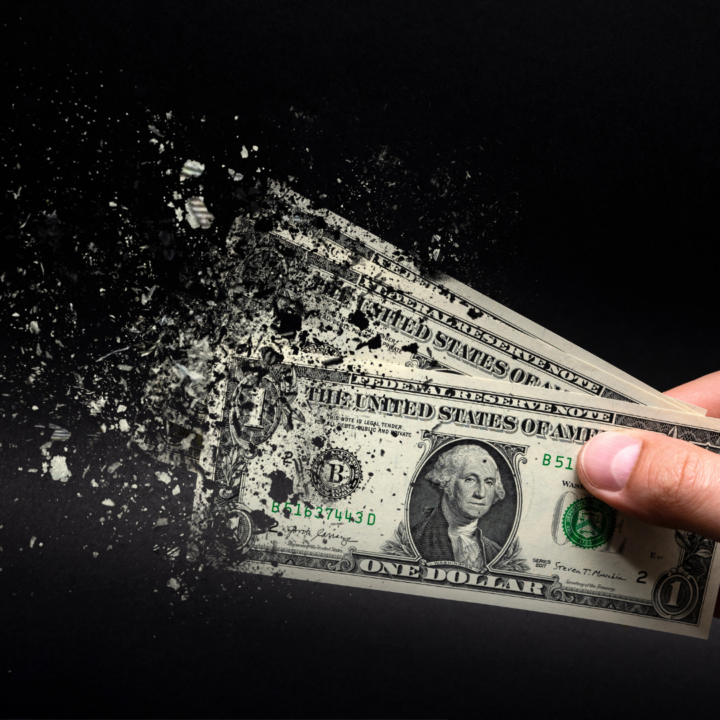 Hand holding US dollar bills that are disintegrating into particles against a black background, symbolizing inflation