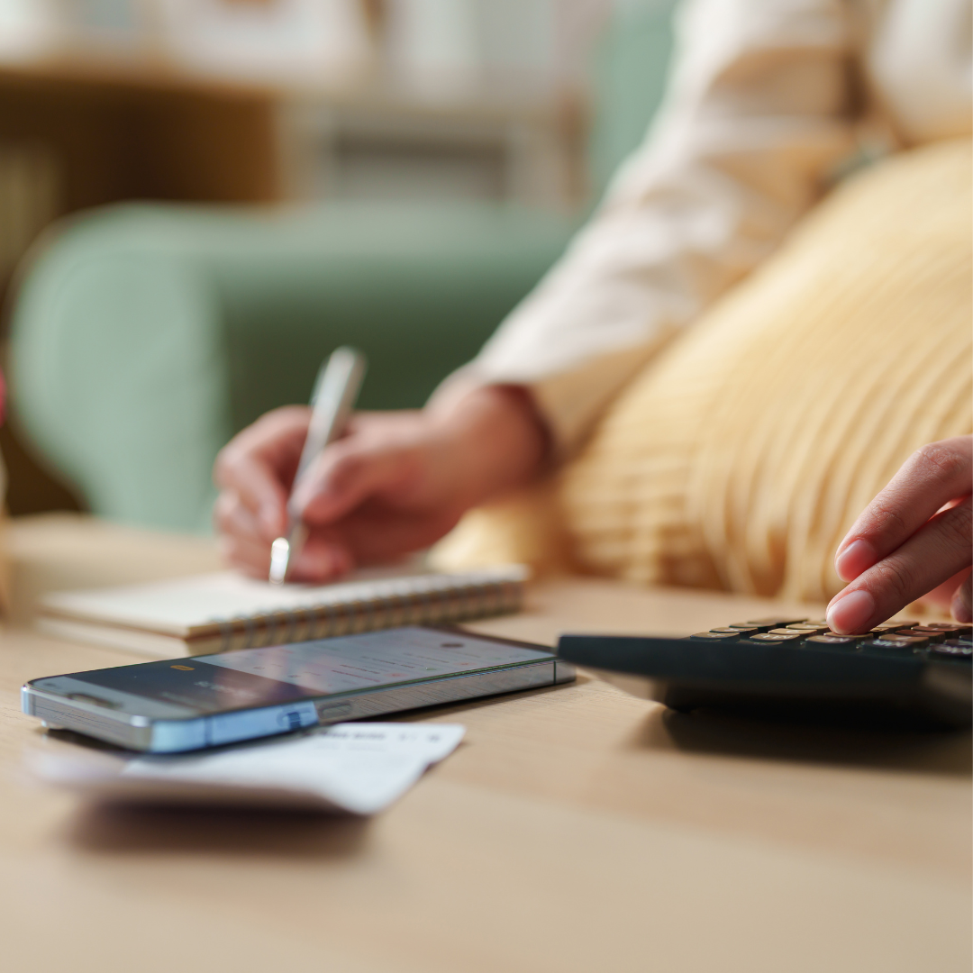 Person using a calculator and writing notes with a smartphone and receipt on the table, managing personal finances